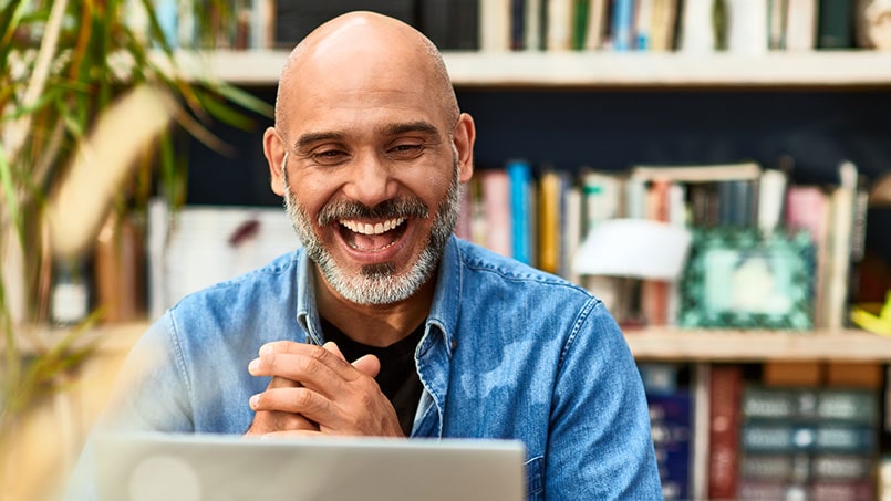 Photo of a middle aged bald man with tanned skin and a short greying beard, wearing a blue denim shirt over a dark t-shirt, looking at a laptop screen and laughing while clapping their hands together. In the background there are shelves with a number of books, photographs in frames and other homely items.