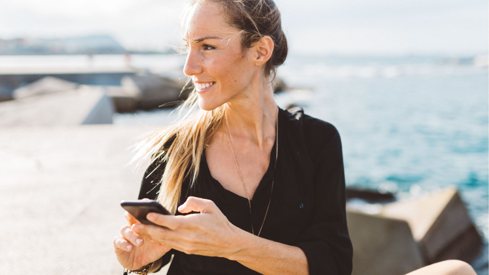 Overcoming Personalisation Paralysis webinar - Photo of a woman with long blonde hair on the beach, wearing a black top and holding a smartphone