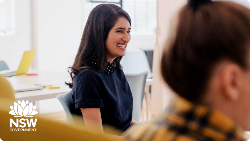 Side-on photo of a woman with long dark hair, wearing a dark blue short-sleeved top. She is laughing. There is another woman in the foreground but only the back of her head is in view and it is our of focus. In the bottom left corner is the NSW government Waratah logo.