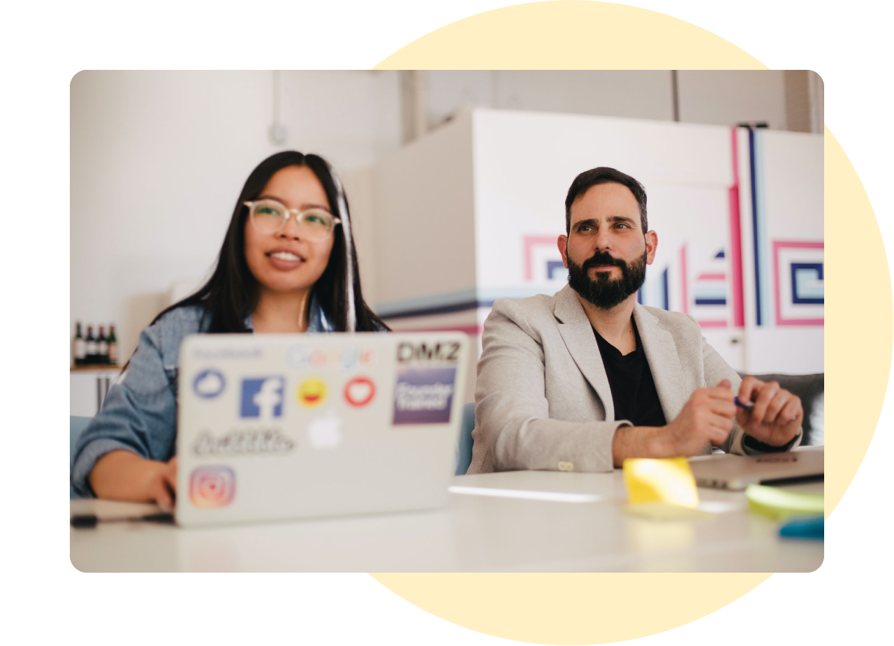 Photo of a man sitting next to a woman at a desk. The man is wearing a cream coloured blazer over a dark t-shirt and has short dark hair with a groomed beard. The woman is wearing glasses and has long dark hair with one streak of white running through it. In front of them is a laptop covered in stickers.