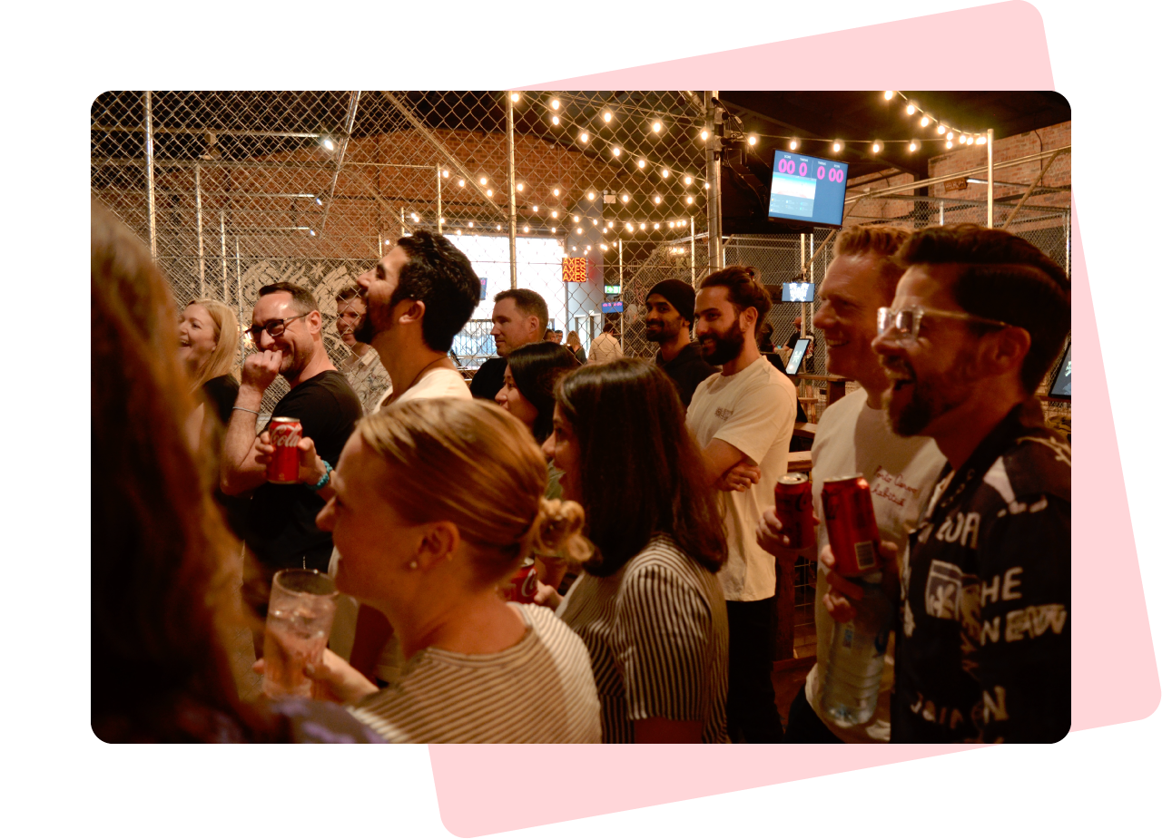 Photo of a group of people at an indoor social gathering. They are all laughing and holding drinks. Overhead, fairy lights are hanging from the ceiling.