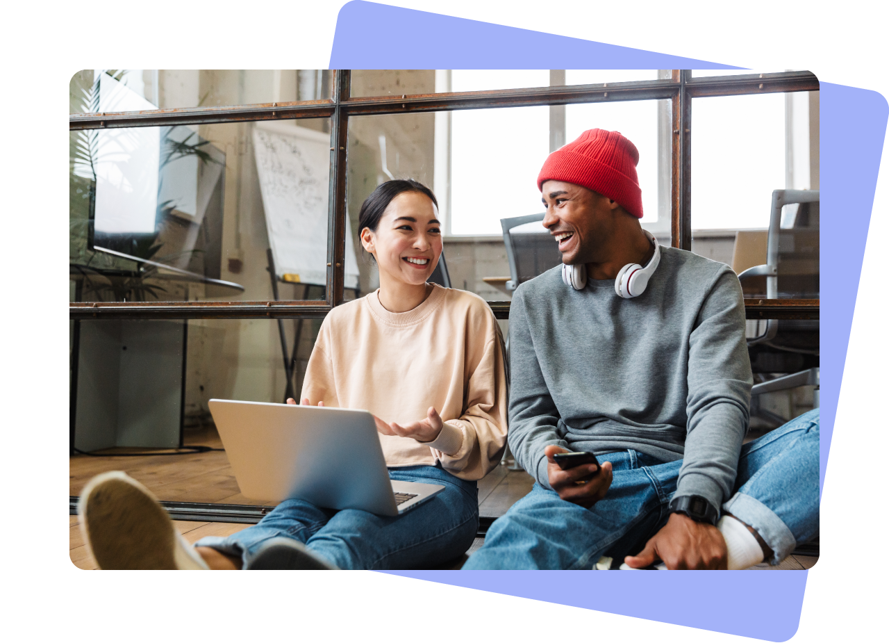 Man and woman sat next to each other on the floor. The man has a red wooly hat, a great jumper and blue jeans. He has white headphones around his neck. The woman has a light pink jumper and blue jeans. She has a laptop on her lap. They are laughing and smiling together.