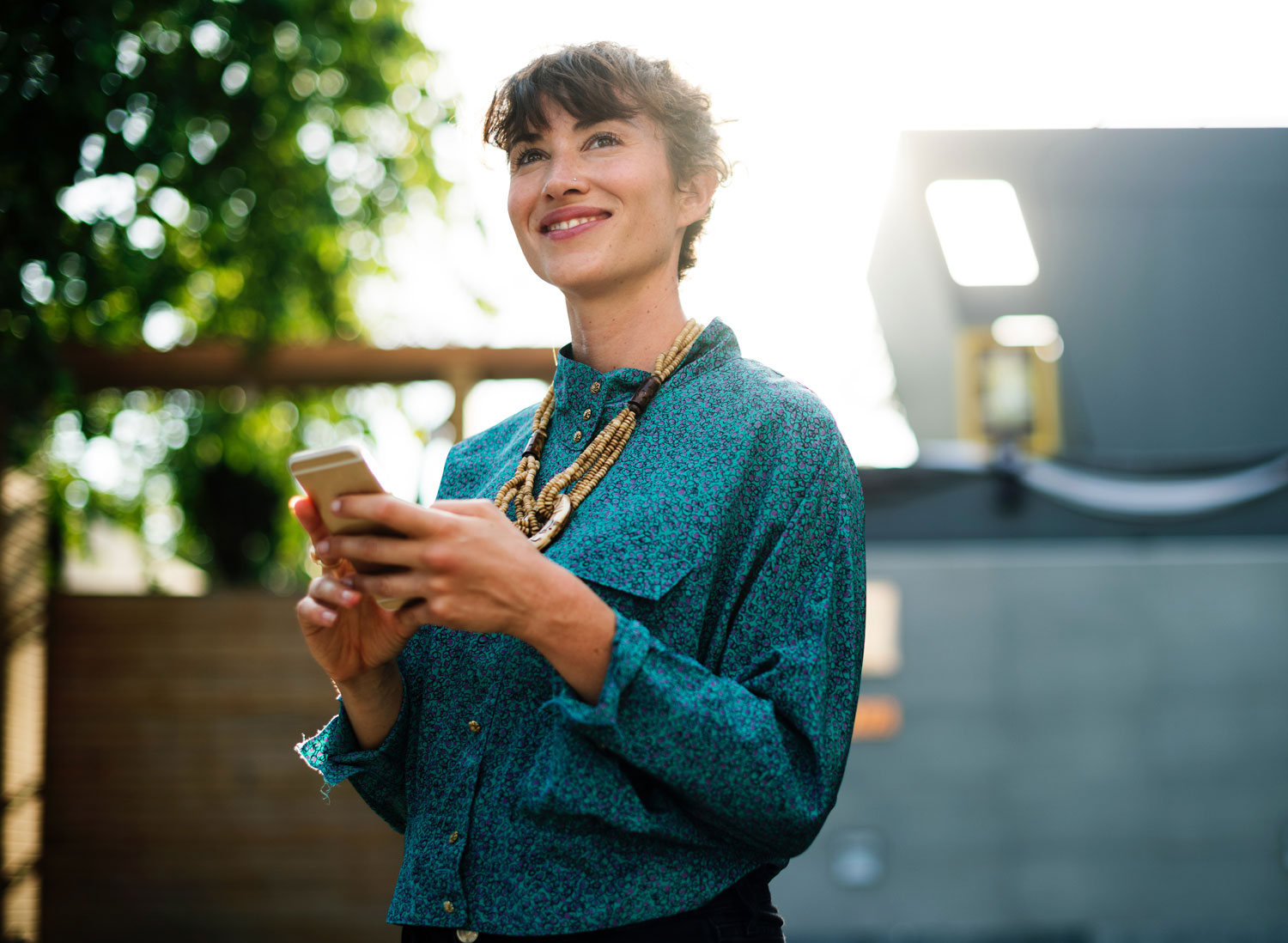 A woman with brown hair tied up behind her head, wearing a green blouse and large necklace is using a mobile device outside. She looks like it is good fun.