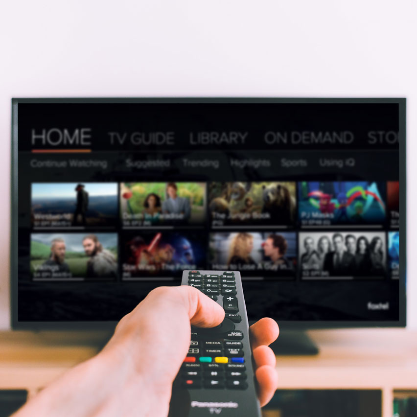 A hand is outstretched in from of the camera, holding a remote control and pointing it at a TV showing a video streaming service.