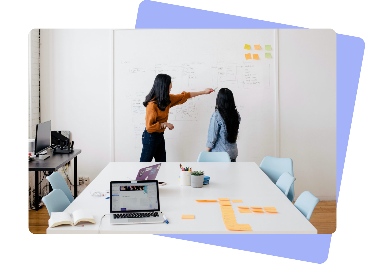 Photo of two women in discussion in front of a whiteboard that is covered in writing, illustrations and post-it notes. The woman on the left is wearing an orange jumper and pointing at something on the whiteboard.