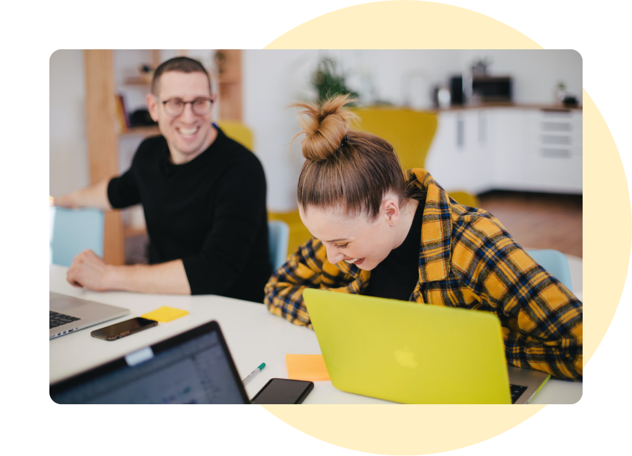 Photo of a man and woman sat together at a desk. The man has very short hair and is wearing a black jumper and glasses. The woman has her hair tied up in a bun and is wearing a yellow tartan jacket. On top of the desk is an open yellow laptop. They are both laughing and the woman is leaning forward.