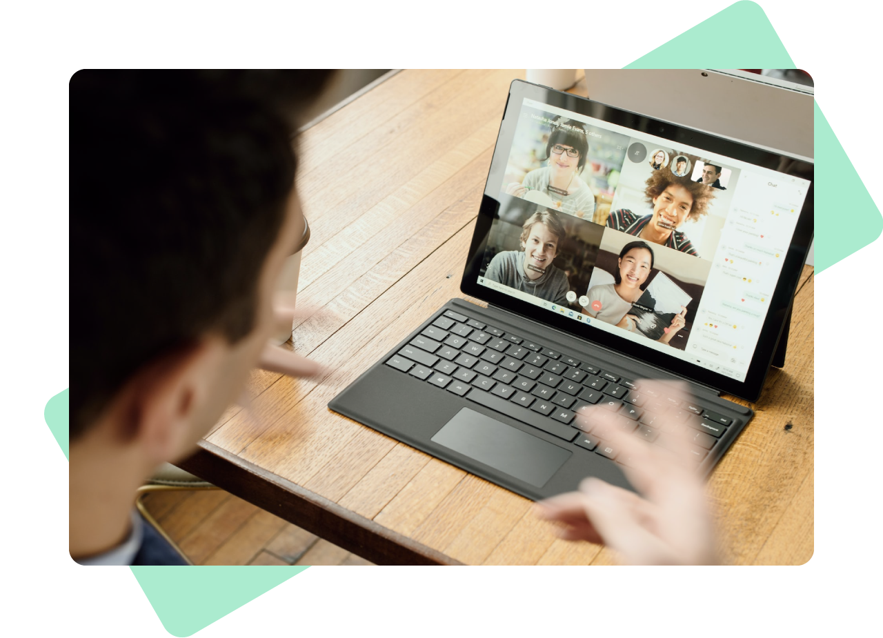 Man looking down at his laptop on a wooden table. On the screen is a video call with four other people.