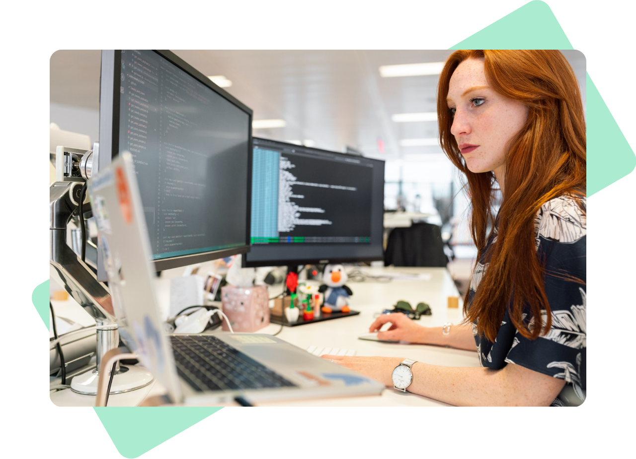 Technical development - photo of a female web developer working at her desk. In front of her is a laptop and two external displays, all showing lines of software code. The desk is covered in lots of personal items.