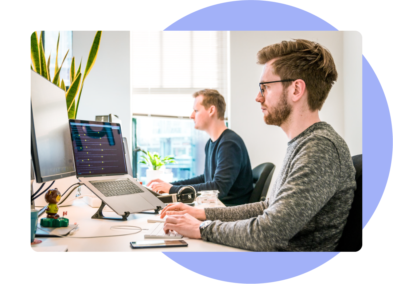 Website support & optimisation - photo of two male web developers at their desk, typing code onto their laptops. In the background there is a window. The desk is covered in plants and computer peripherals.