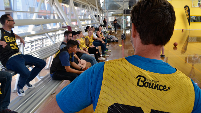 Over the shoulder photograph of a group of men and women inside a sports hall. The person in front of the camera is wearing a yellow Charity Bounce sports vest over a blue t-shirt and holding a basketball under their arm.