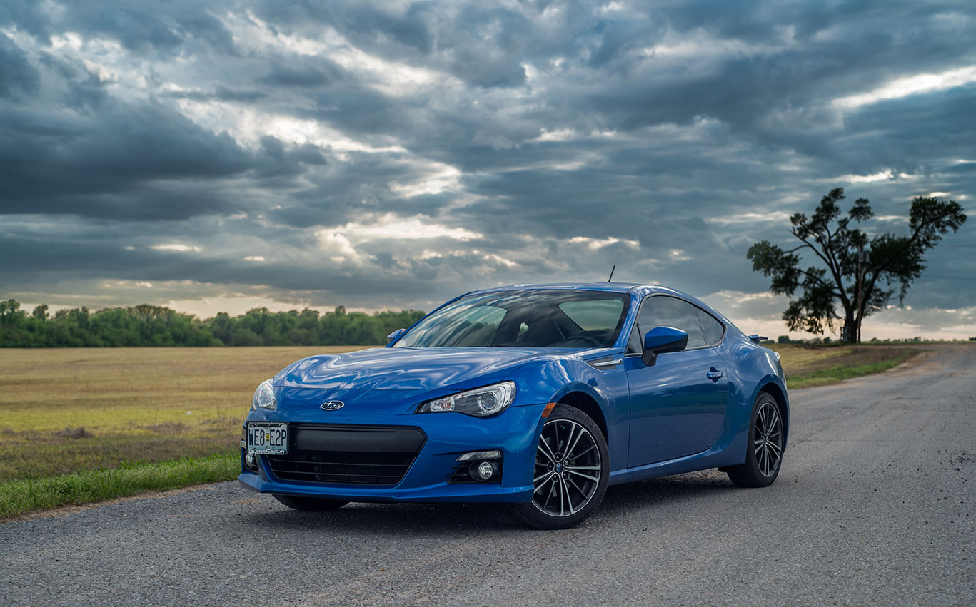 Blue Subaru BRX parked on a gravelly road in front of a field flanked by woods. The sky overhead is cloudy and moody.