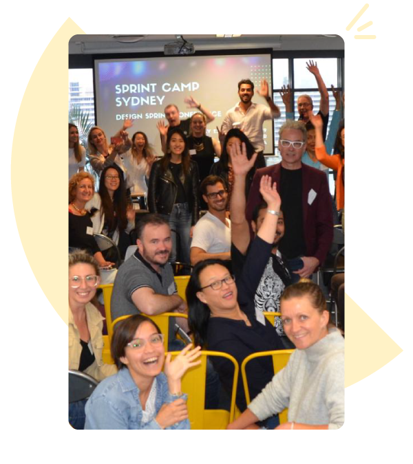 Photo of a room full of people attending a Sprint Camp event. They are all turning around and waving to the camera. In the background there is a projector screen.