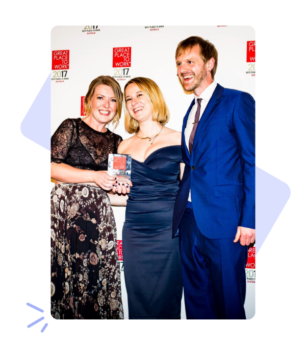 Photo of one man and two women posing in front of a Great Place To Work awards background. They are all wearing formal clothing and smiling.