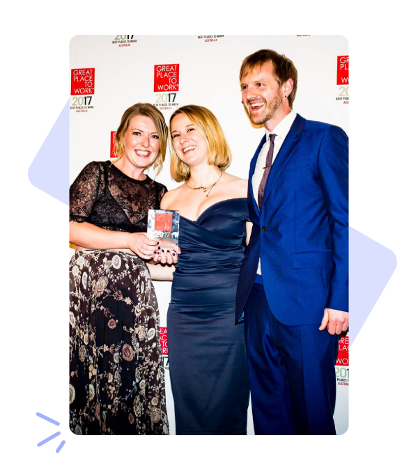 Photo of one man and two women posing in front of a Great Place To Work awards background. They are all wearing formal clothing and smiling.