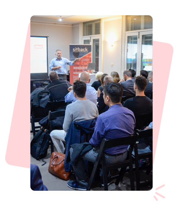 Photo of a room full of different people sat in rows, listening to a man present in front of a projector screen.