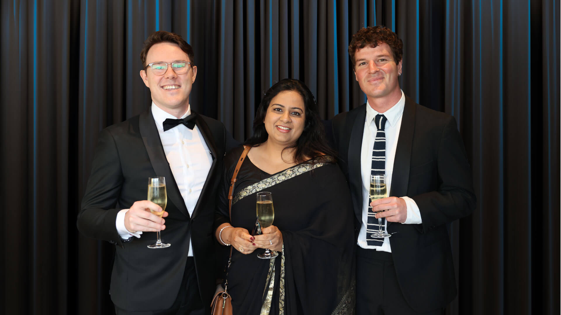 The winning Sydney Opera House team stood in front of a curtain at the Acquia Engage Awards 2023. They are dressed in formal black tie and holding champagne glasses.