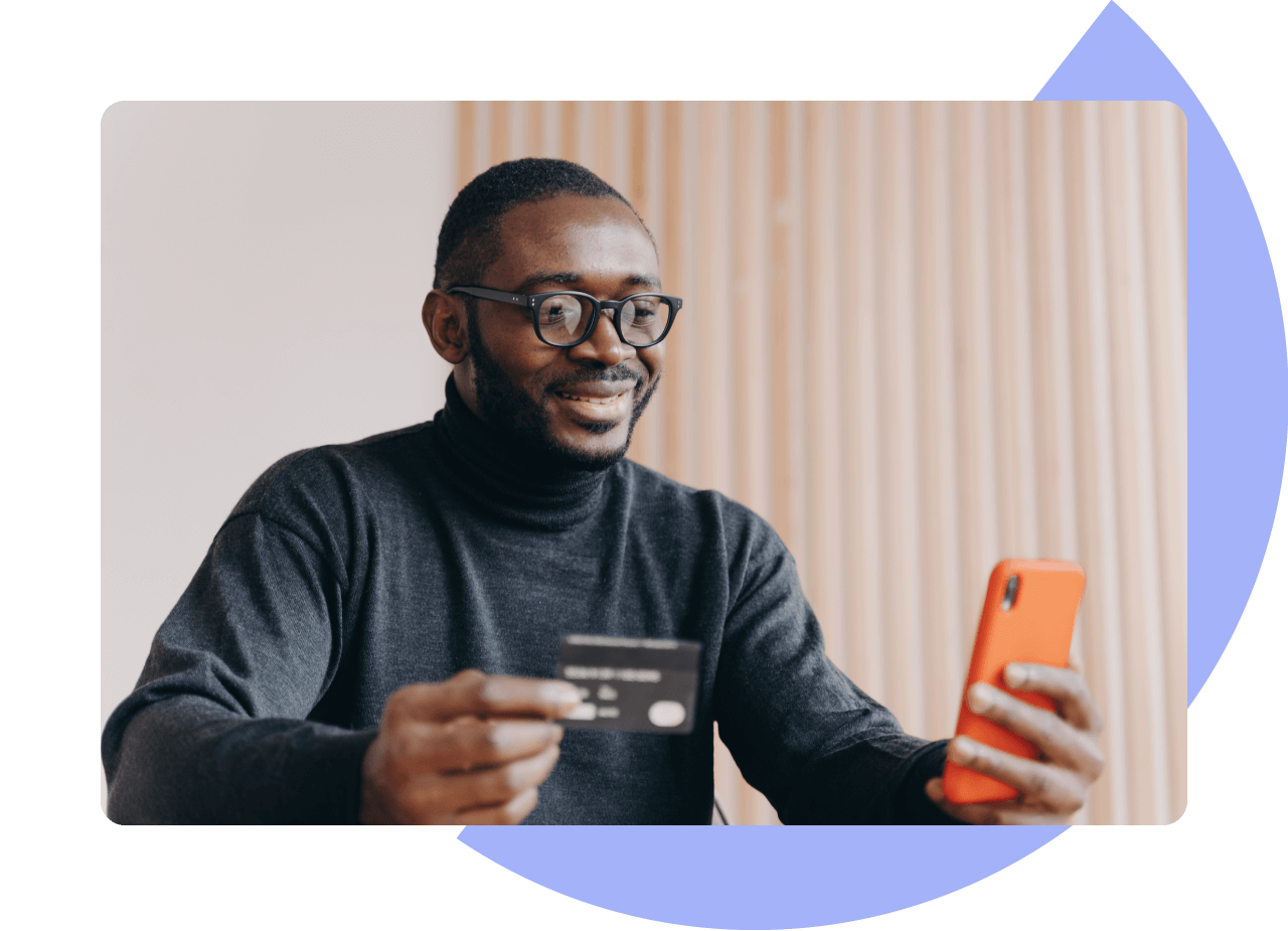 Man sat at a table looking at his mobile phone while holding a credit card.