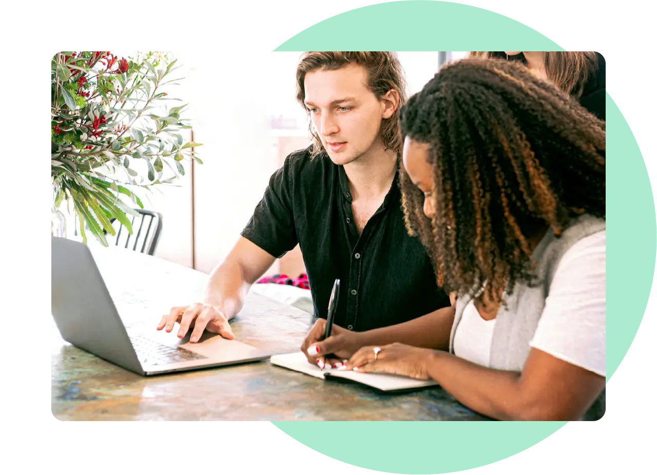 Two colleagues working together at a laptop while taking notes during a collaborative content planning session.