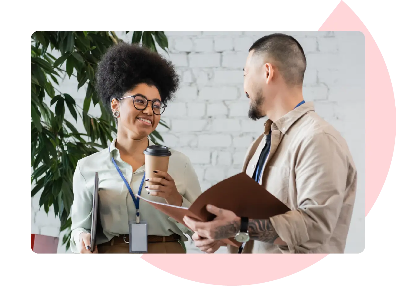 Two coworkers smiling while having a relaxed conversation with coffee and notebooks in hand.