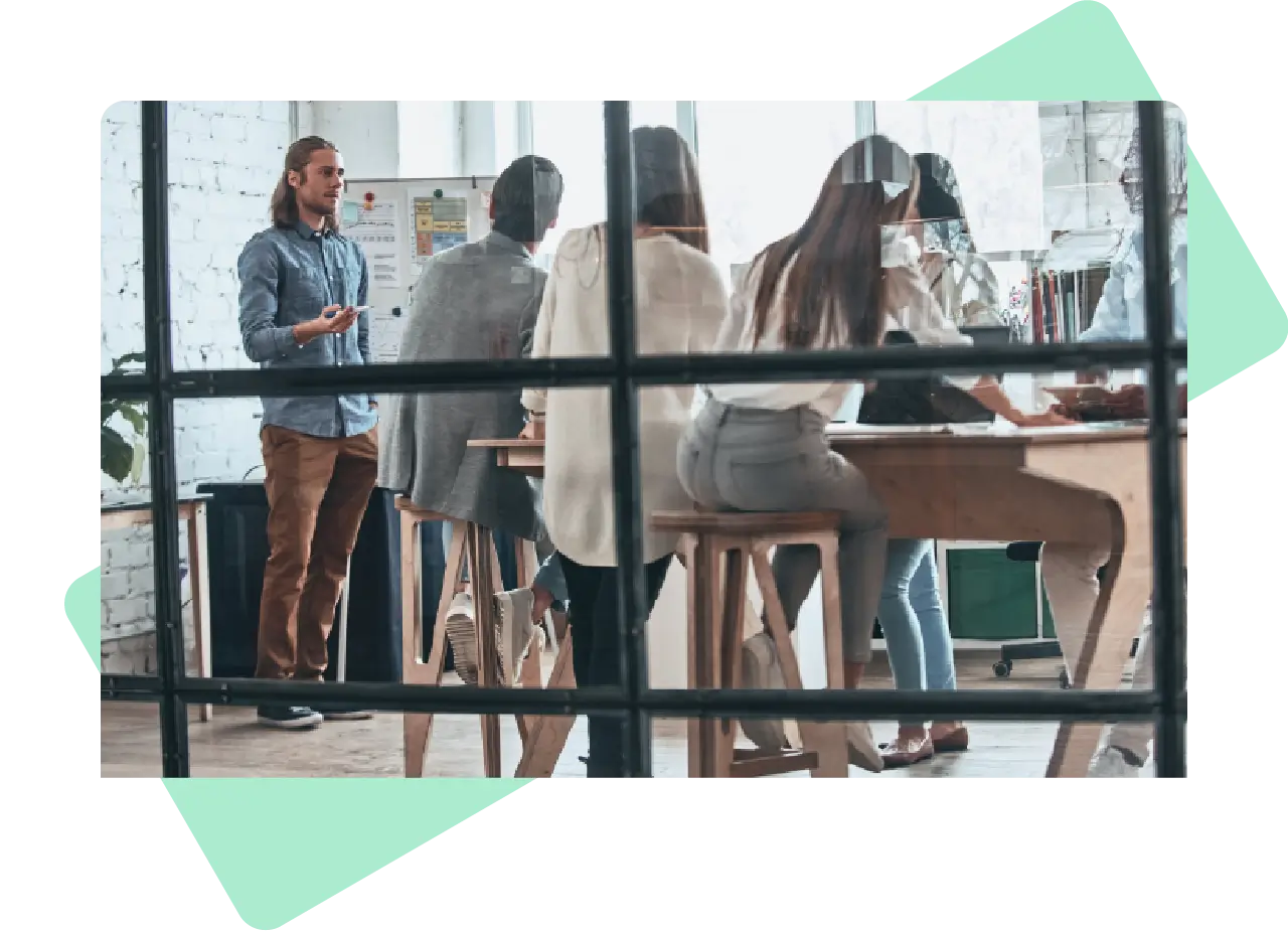 View through glass of a team in a collaborative meeting, with one person presenting to a group seated around a high table in a modern office.