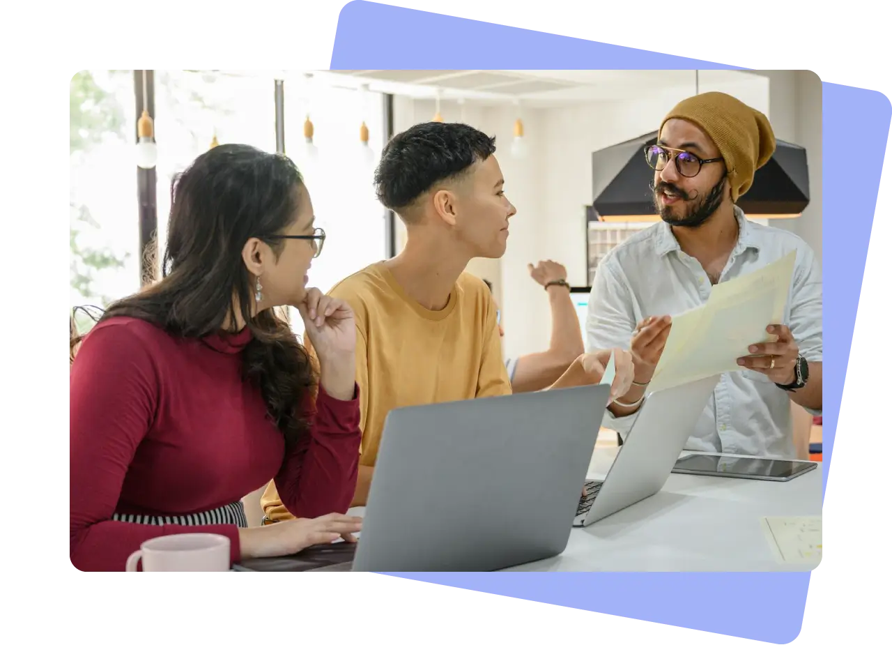 Three colleagues collaborating around a desk with laptops and printed documents, discussing content ideas in a bright modern workspace.