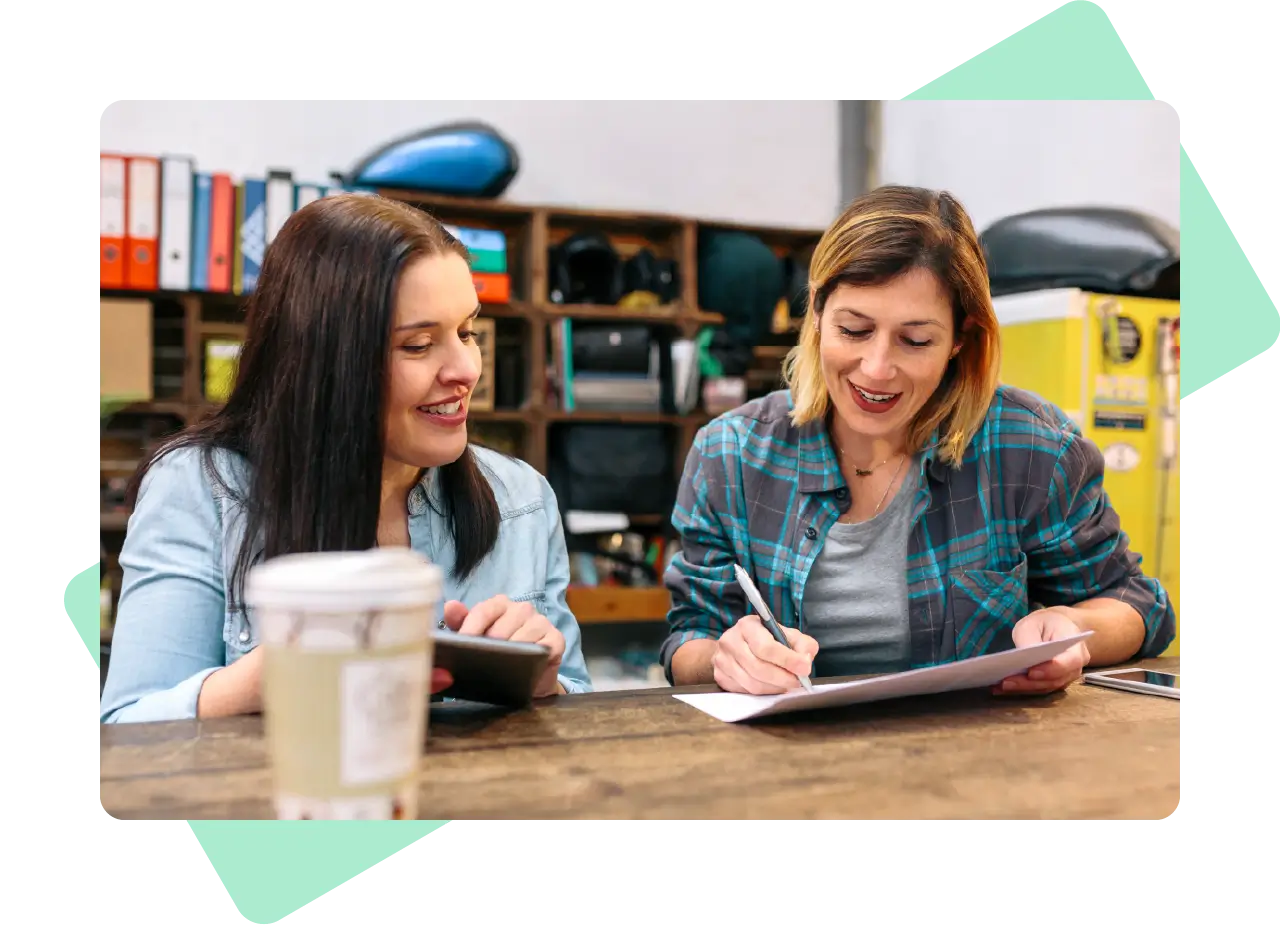 Two women collaborating at a rustic table, reviewing notes and sharing ideas with a tablet and paper in front of them.