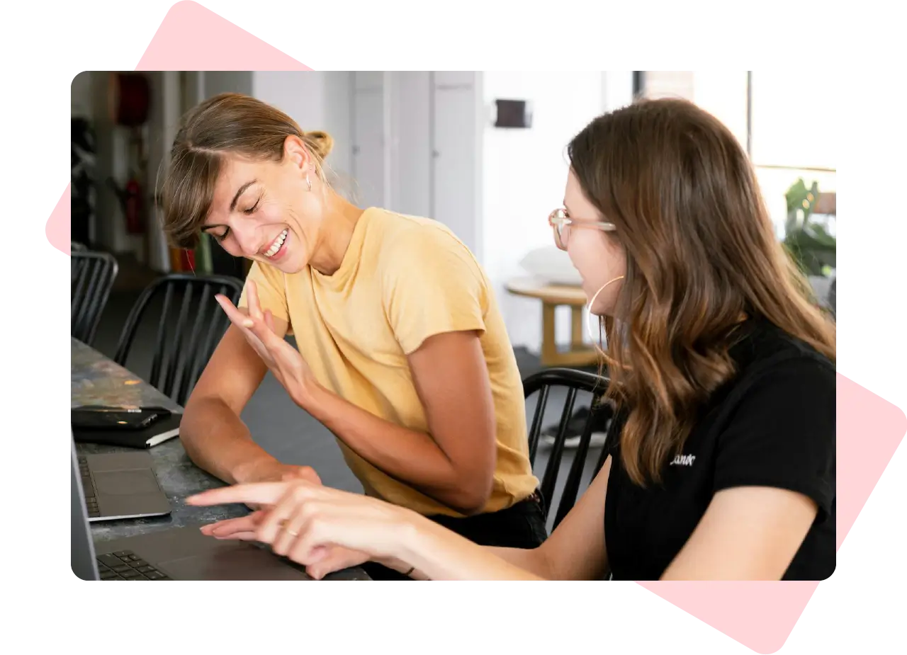 Two women laughing together during a content review session at a laptop in a casual workspace.